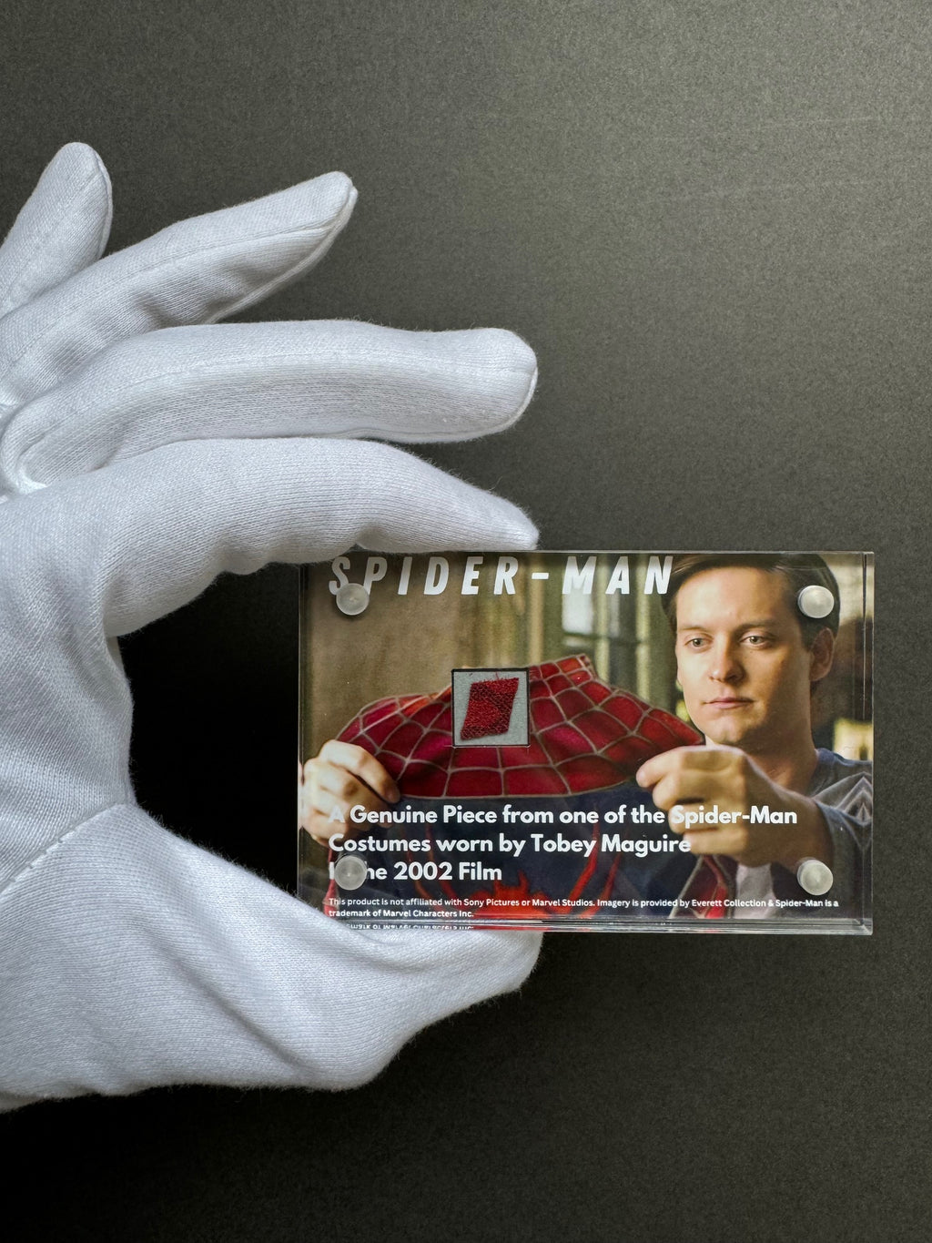A gloved hand presenting a miniature movie prop display case with an image of Tobey Maguire in the Spider-Man costume. 